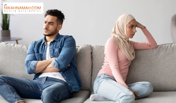 A Muslim couple sitting on a couch facing away from each other, appearing upset. The man is wearing a denim jacket and has his arms crossed, while the woman, wearing a hijab and a pink sweater, rests her head on her hand. The Nikahnamah.com logo is visible in the top left corner, representing a Muslim matrimonial platform